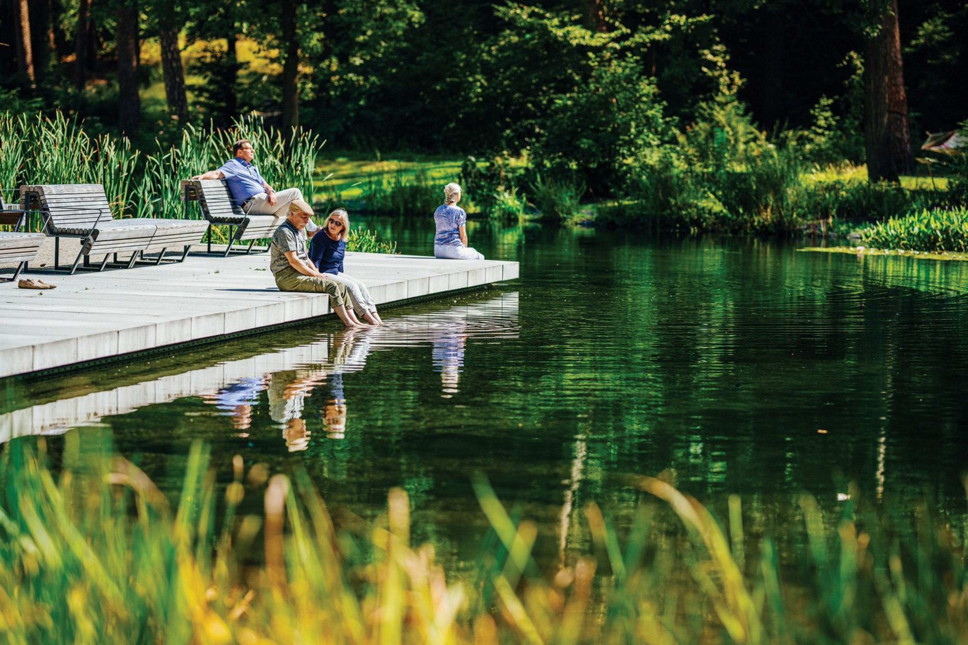 Auf einem Steg an einem See sitzen vier Personen. drei halten ihre Füße ins Wasser. Im Hintergrund sind Bäume zu sehen.