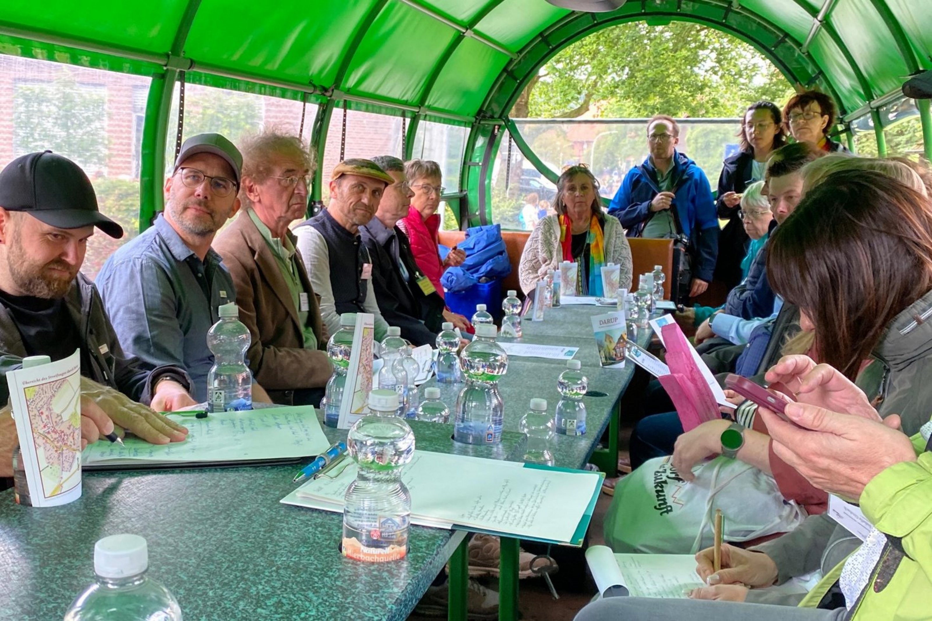 Siebzehn Personen sitzen in einem Planwagen mit grüner Plane an einem Tisch. Vor ihnen stehen Getränke und manche schauen in Broschüren.