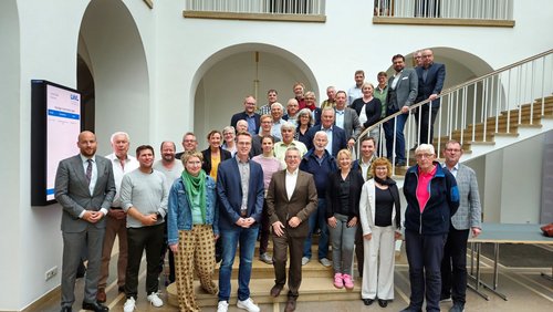 Die Mitglieder der Verbandsversammlung stehen in einem hellen Treppenhaus. 