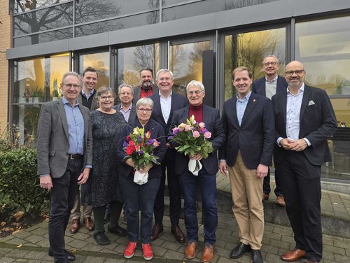 Elf Personen stehen in zwei bis drei Reihen hintereinander. Eine Frau und ein Mann halten jeweils einen Blumenstrauß in der Hand. Hinter der Gruppe befindet sich eine Fensterfront eines Gebäudes. 