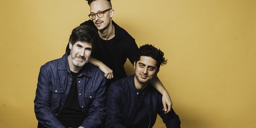 Drei Männer sitzen vor einem gelben Hintergrund. Zwei sitzen nebeneinander, der Dritte hockt hinter ihnen und legt seine Arme auf deren Schultern.