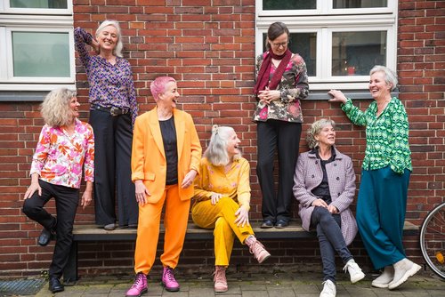 Sieben Frauen stehen und sitzen auf und vor einer Bank, die vor einem roten Backsteinhaus steht. Die Frauen tragen bunte Kleidung und lachen.