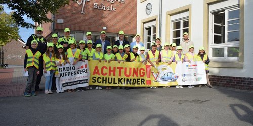 Eine Gruppe von Kindern und Erwachsenen in gelben Warnwesten und gelben Mützen hält einen Banner mit der Aufschrift Achtung Schulkinder