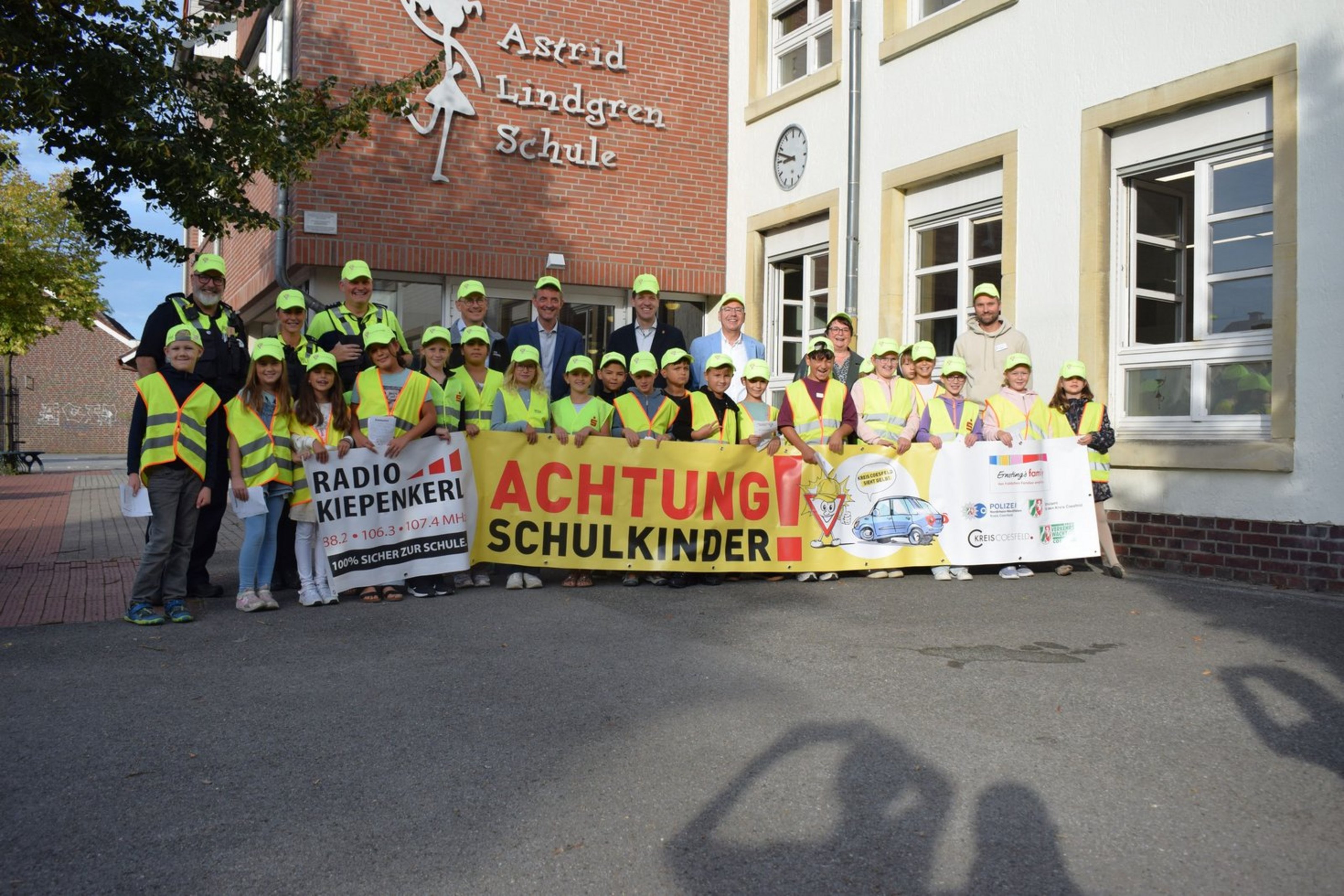 Eine Gruppe von Kindern und Erwachsenen in gelben Warnwesten und gelben Mützen hält einen Banner mit der Aufschrift Achtung Schulkinder