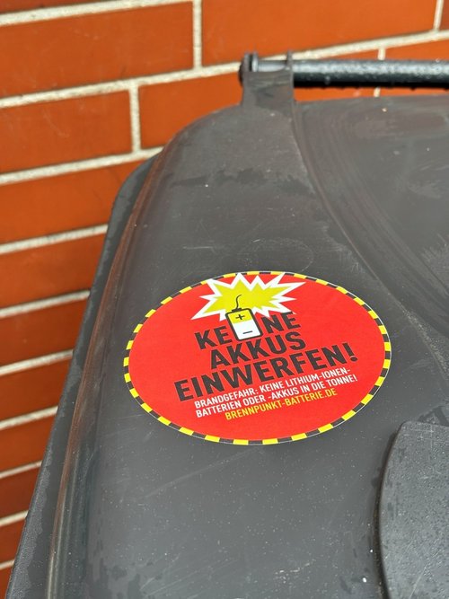 Man sieht den Deckel einer Mülltonne mit einem roten Sticker, auf dem Keine Akkus einwerfen steht.