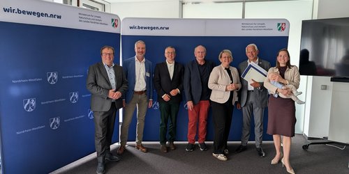 Sieben Personen stehen vor einer blauen Wand, auf der "wir.bewegen.nrw." steht.