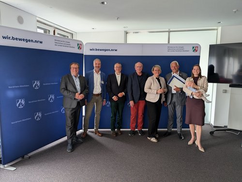 Sieben Personen stehen vor einer blauen Wand, auf der "wir.bewegen.nrw." steht.