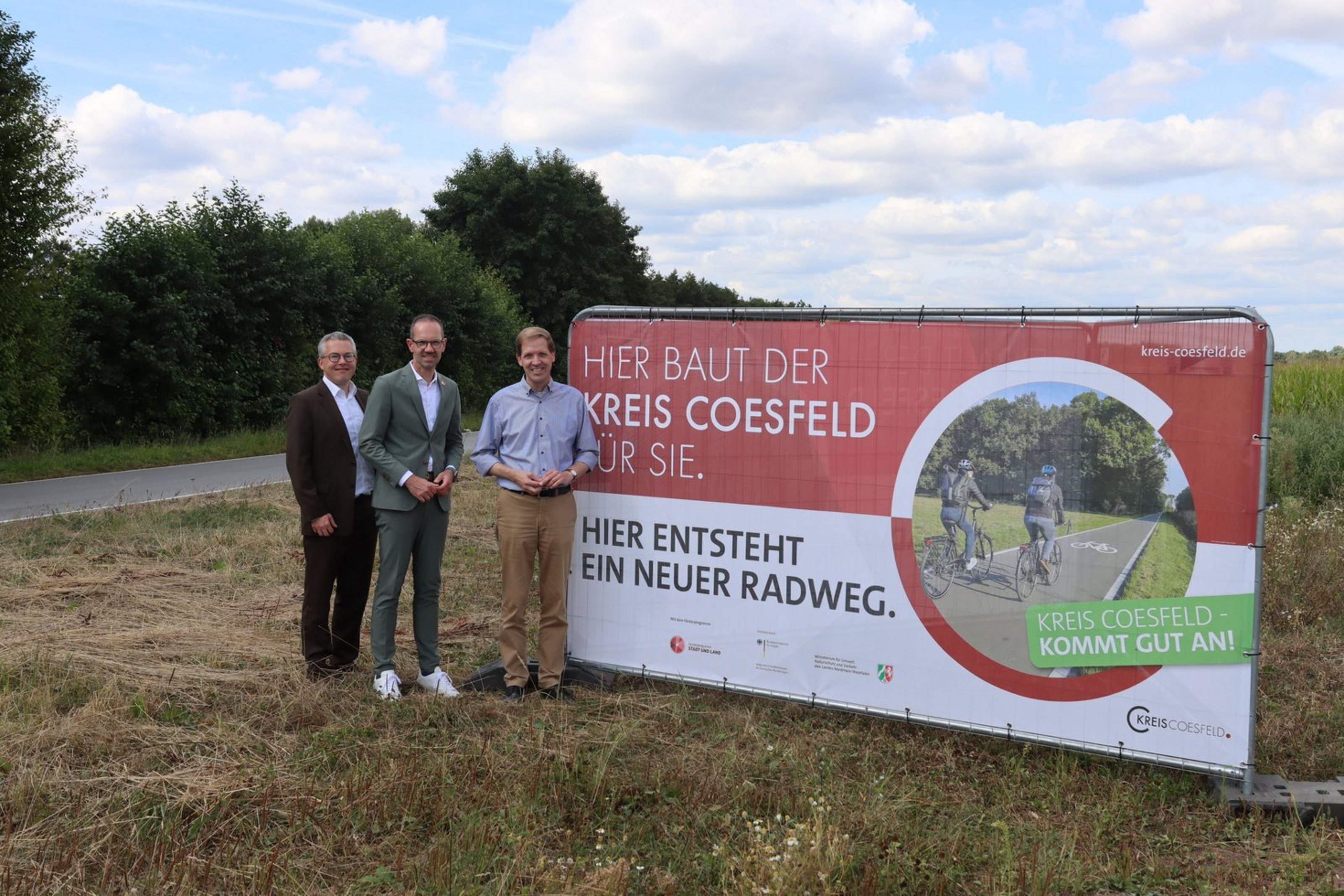 Kreisdirektor Dr. Linus Tepe, Bürgermeister Ansgar Mertens, Stadt Lüdinghausen und Landrat Dr. Christian Schulze Pellengahr an der K16 (Foto: Kreis Coesfeld). Am Straßenrand ist ein Banner aufgebaut, auf dem "HIer baut der Kreis Coesfeld für Sie" steht. Darunter steht "Hier entsteht ein neuer Radweg". Links vom Schild stehen drei Männer in förmlicher Kleidung und schauen in die Kamera.
