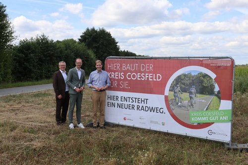 Am Straßenrand ist ein Banner aufgebaut, auf dem "HIer baut der Kreis Coesfeld für Sie" steht. Darunter steht "Hier entsteht ein neuer Radweg". Links vom Schild stehen drei Männer in förmlicher Kleidung und schauen in die Kamera.
