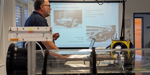 Ein Mann steht in einem Klassenraum. An der Wand sind zwei Autos abgebildet. Im Vordergrund ist eine durchsichtige Röhre auf einem Gestell befestigt.