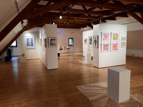 Blick in eine Kunstausstellung mit Gemälden und Skulpturen in einem hellen Raum mit Holzbalkendecke.