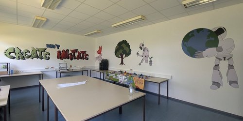In einem Klassenraum steht ein Gruppentisch in der Mitte, am Rand sind weitere Tische zu sehen. An der Wand sind Bilder von Naturmotiven und Robotern zu sehen sowie der Schriftzug "Create don't imitate".