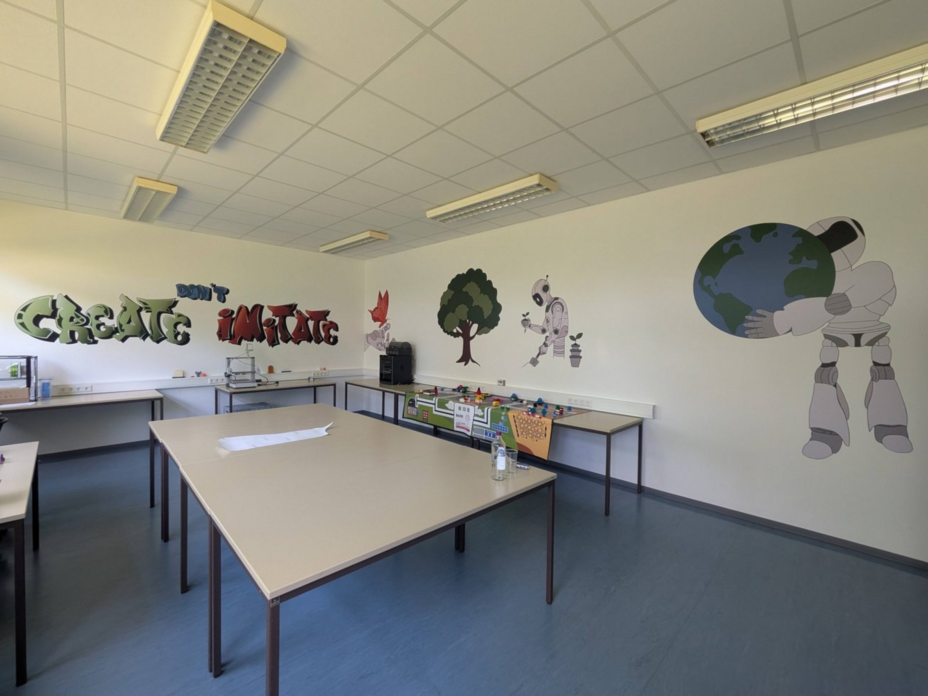 In einem Klassenraum steht ein Gruppentisch in der Mitte, am Rand sind weitere Tische zu sehen. An der Wand sind Bilder von Naturmotiven und Robotern zu sehen sowie der Schriftzug "Create don't imitate".
