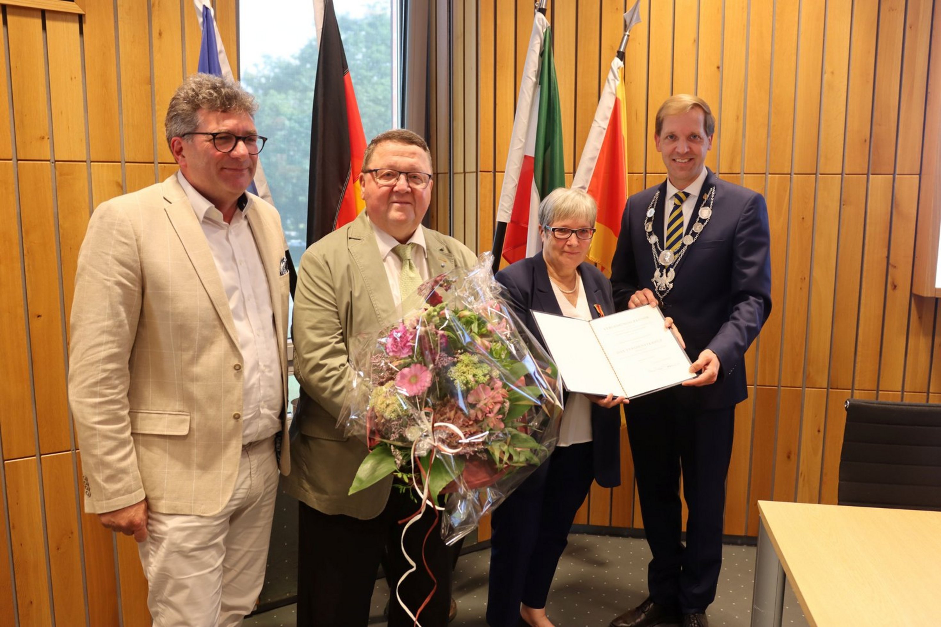 (v.l.n.r.) Jörn Möltgen, Bürgermeister der Gemeinde Havixbeck, Herr Köhler, Lebensgefährte von Margarete Schäpers, Margarete Schäpers und Dr. Christian Schulze Pellengahr (Foto: Kreis Coesfeld). Vier Personen in förmlicher Kleidung stehen nebeneinander vor vier Fahnen. Eine Person hält einen Blumenstrauß, die einzige Frau aus der Gruppe hält eine Urkunde in der Hand.