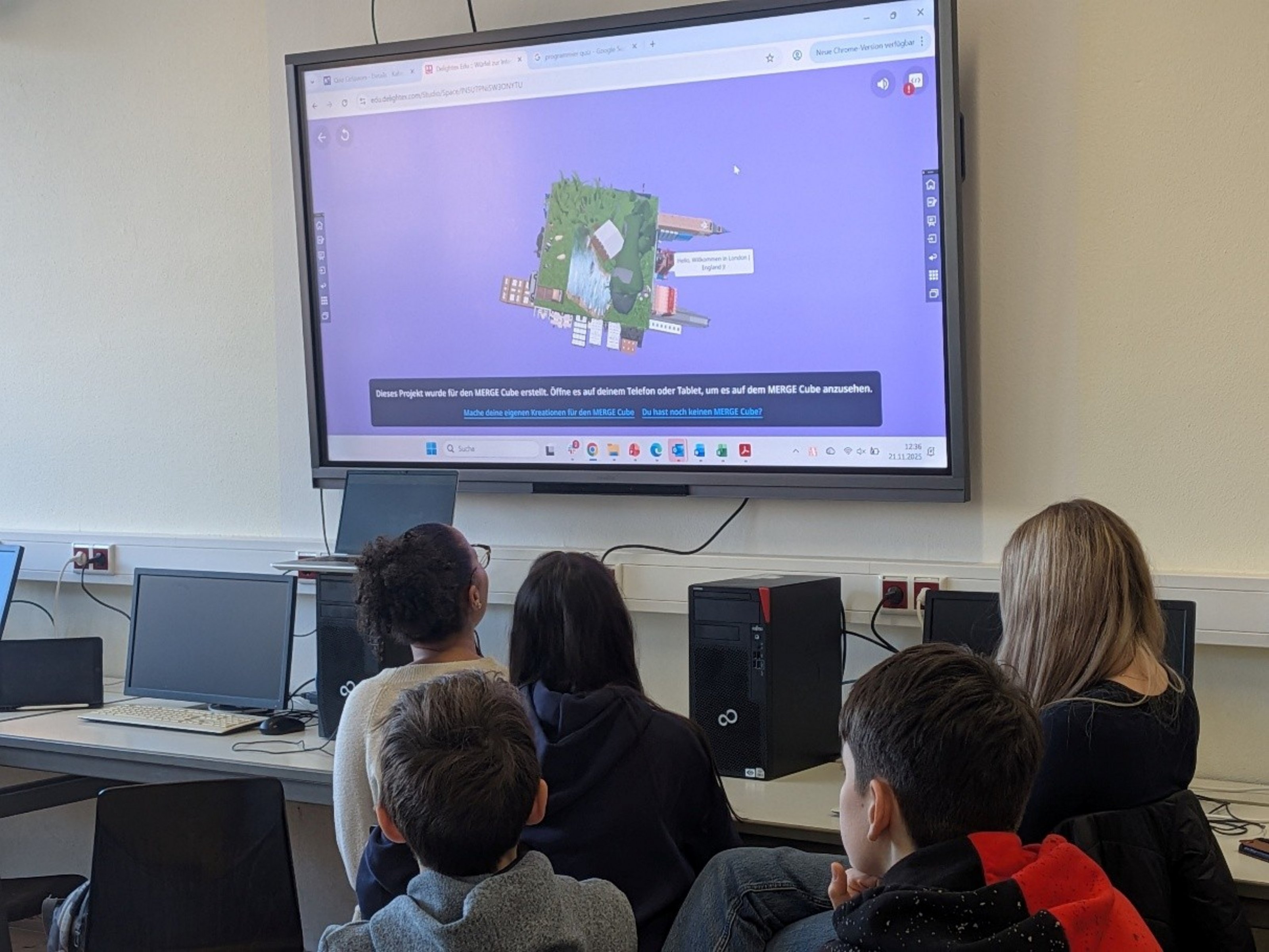 Fünf Kinder schauen auf einen Bildschirm, der über einem Tisch mit Computern hängt. 