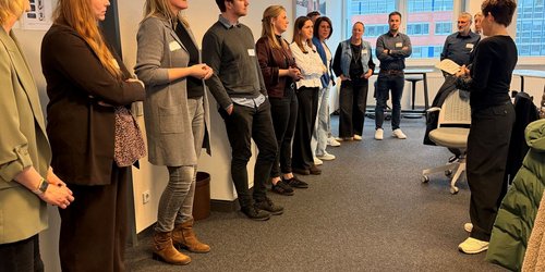 Teilnehmerinnen und Teilnehmer stehen im Halbkreis in einem Besprechungsraum und diskutieren im Workshop über Anwerbung und Integration internationaler Fachkräfte.