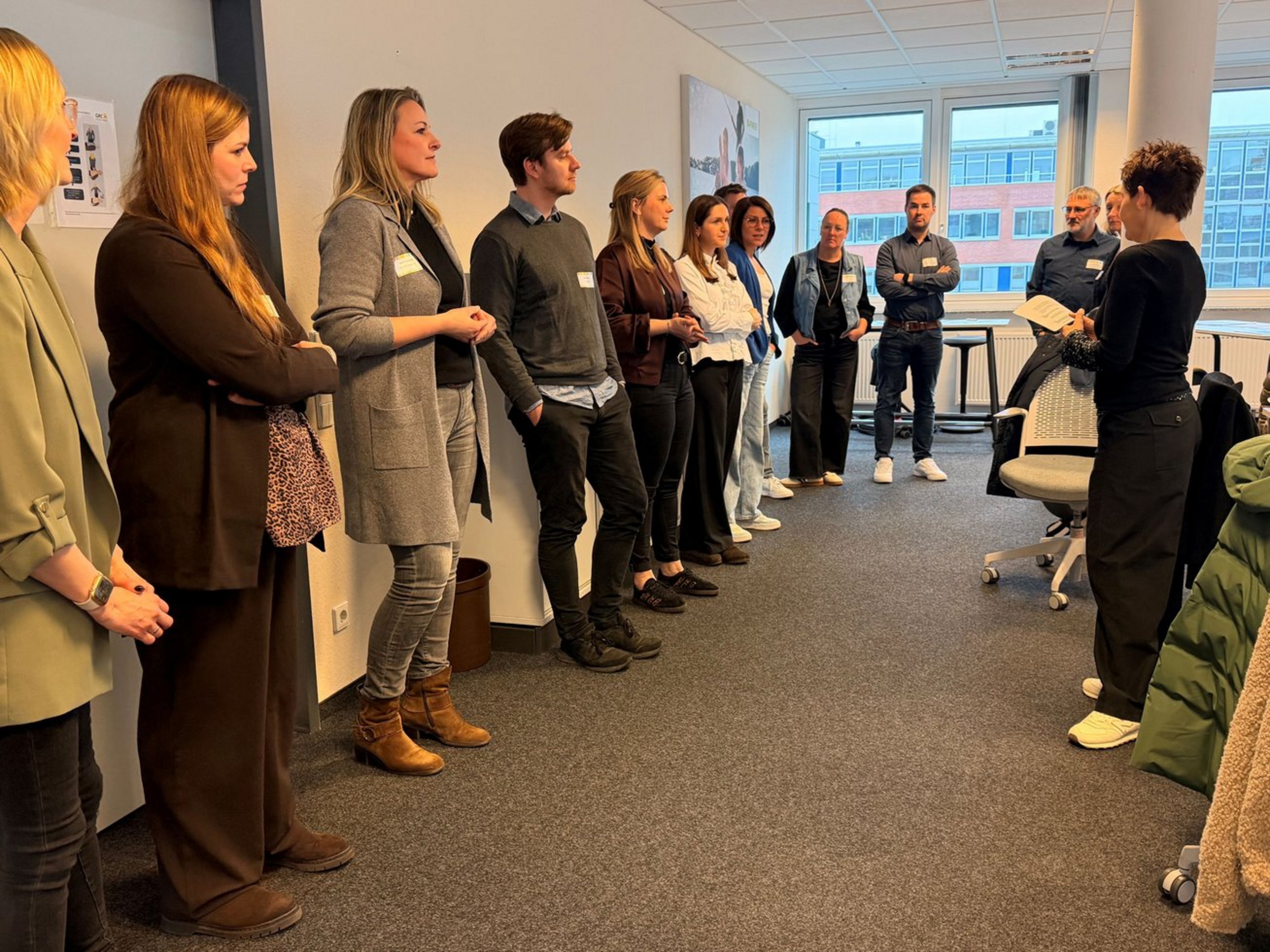 Teilnehmerinnen und Teilnehmer stehen im Halbkreis in einem Besprechungsraum und diskutieren im Workshop über Anwerbung und Integration internationaler Fachkräfte.