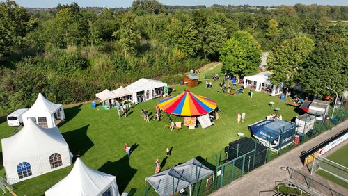 Luftaufnahme eines bunten Festivalgeländes mit Zelten, Ständen und Besuchenden auf einer grünen Wiese.