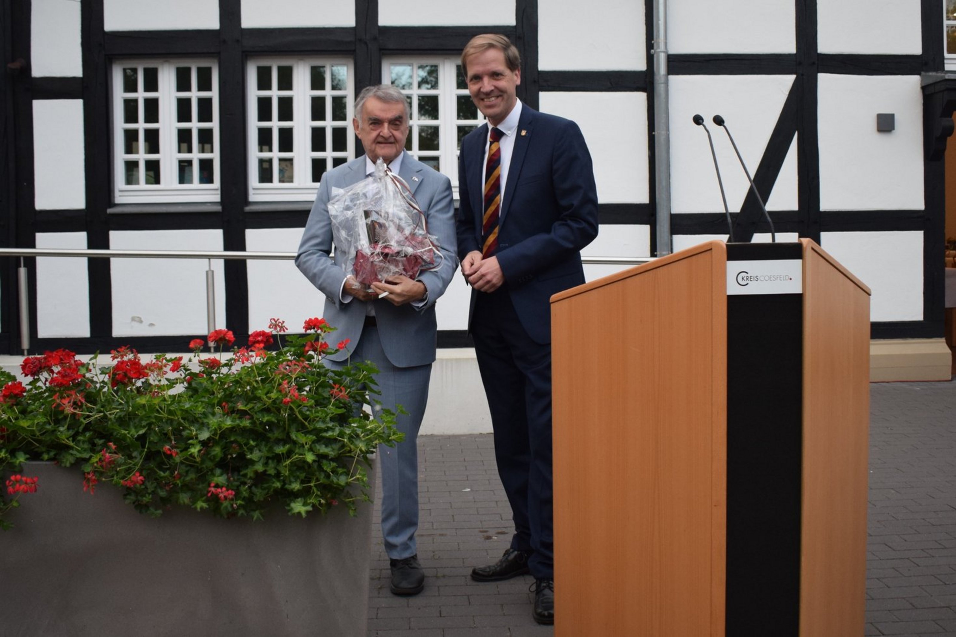 Landrat Dr. Schulze Pellengahr mit Minister Herbert Reul beim Sommerempfang im Garten von St. Lambertus (Foto: Kreis Coesfeld). Hinter einem Rednerpult, das vor einem Fachwerkgebäude steht, stehen zwei Männer in Anzug. Der linke hält ein Präsent in den Händen.