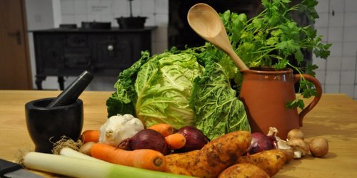 Auf einem Holztisch, der in einer Küche steht liegenverschiedene Gemüse wie Lauch, Möhre, Kartoffel und Zwiebel.