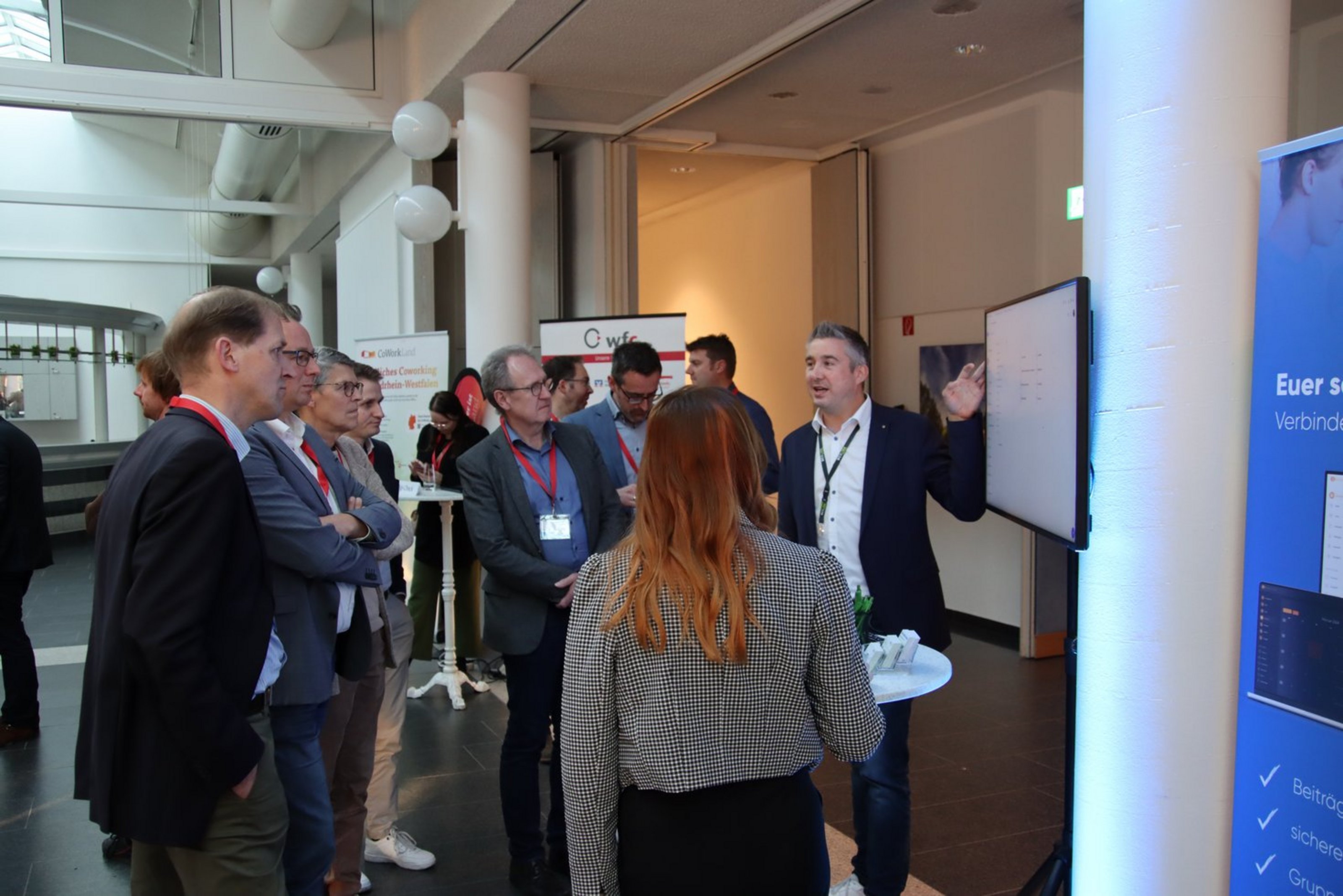Gruppe von Personen mit Namensschildern steht vor einem Bildschirm in einem modernen Konferenzraum und hört einem Mann zu, der auf den Bildschirm zeigt