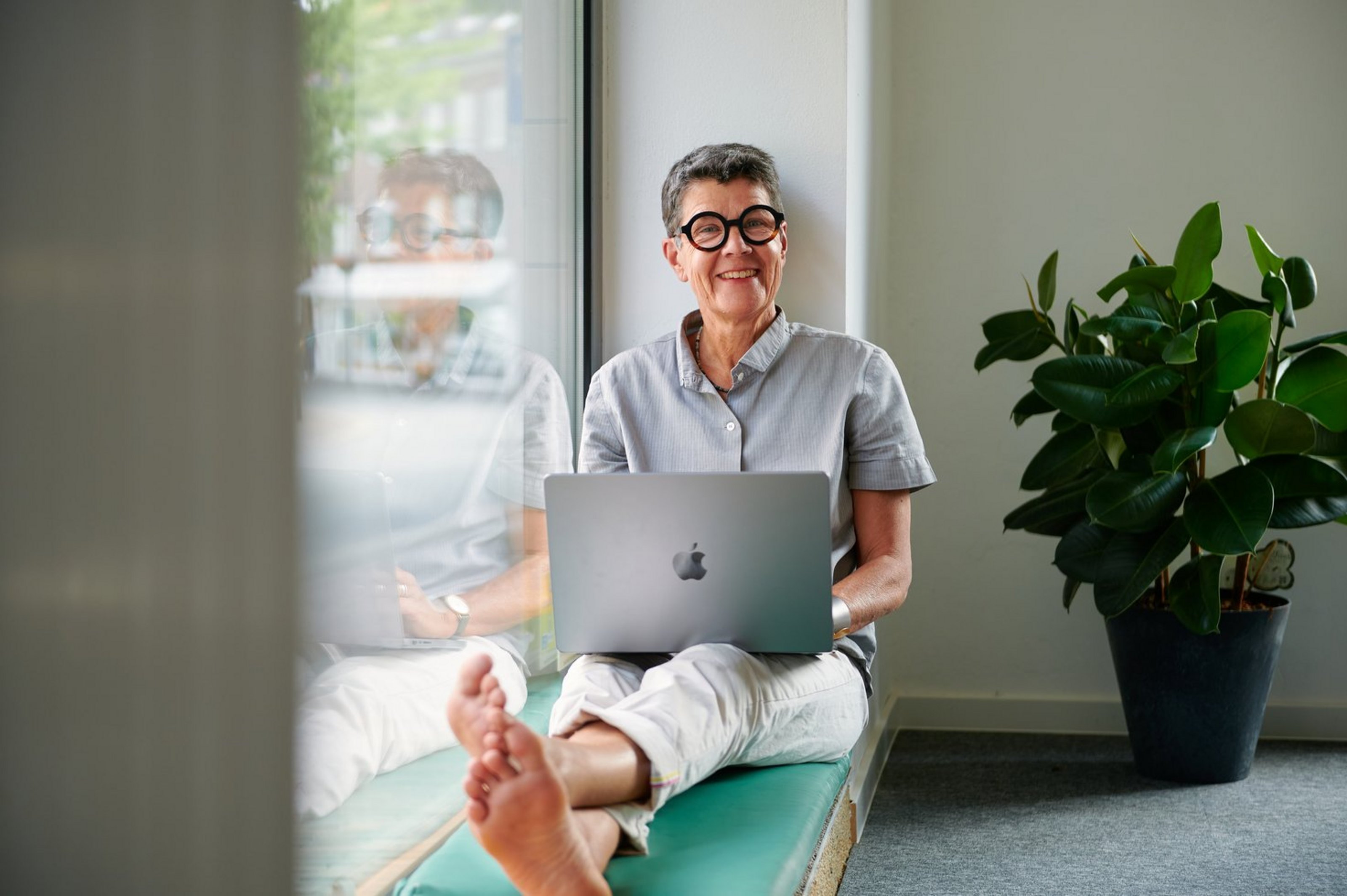 Person sitzt entspannt lächelnd mit einem Laptop auf dem Schoß in einem hellen Innenraum auf einer flachen Fensterbank mit Pflanze im Hintergrund.