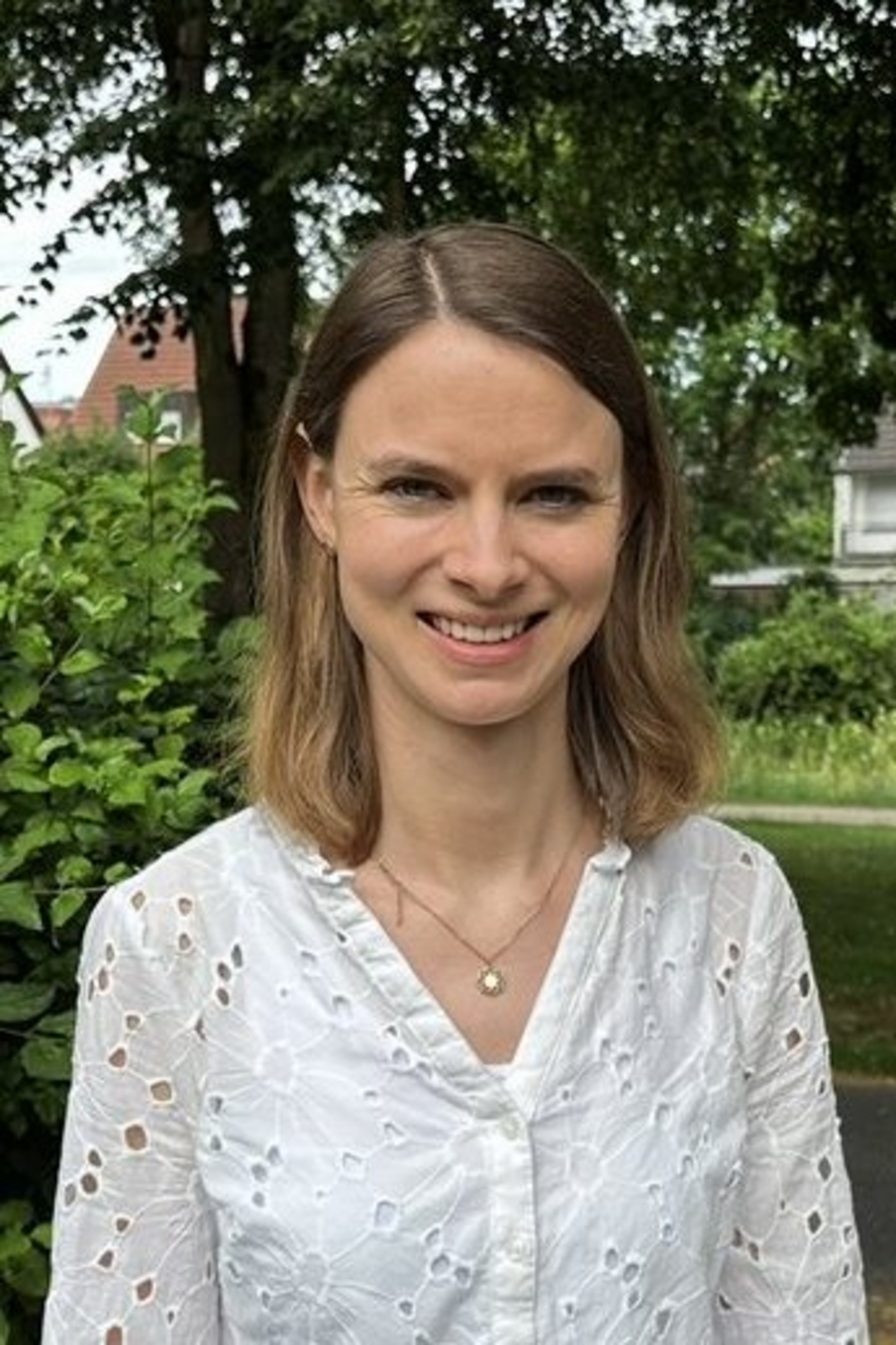 Ein Porträtbild mit der Ansprechpartnerin Sonja Treus: Sie trägt eine dunkle Bluse und steht vor einigen Bäumen mit einer Hecke.
