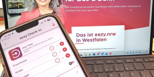 Ein Smartphone wird vor einem geöffneten Laptop gehalten. Auf dem Smartphone ist die App ‚eezy Check-In‘ zu sehen, mit der Fahrgäste für Bus und Bahn einchecken können. Im Hintergrund zeigt der Laptop die Website von eezy.nrw mit der Überschrift ‚Der elektronische Tarif für Bus & Bahn‘. Daneben ist das Foto einer lächelnden älteren Frau abgebildet.
