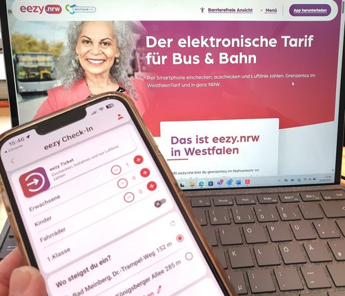 Ein Smartphone wird vor einem geöffneten Laptop gehalten. Auf dem Smartphone ist die App ‚eezy Check-In‘ zu sehen, mit der Fahrgäste für Bus und Bahn einchecken können. Im Hintergrund zeigt der Laptop die Website von eezy.nrw mit der Überschrift ‚Der elektronische Tarif für Bus & Bahn‘. Daneben ist das Foto einer lächelnden älteren Frau abgebildet.