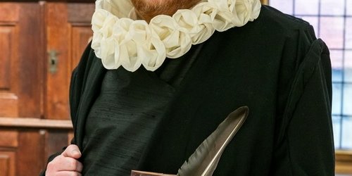 Ein Mann in historischem, schwarezn Gewand mit weißer Halskrause und Hut steht vor einem Holzschrank und hält ein Buch und eine Feder in der Hand.