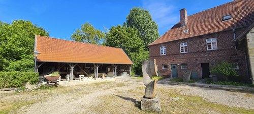 Ein sonniger Innenhof mit zwei Gebäuden. Links steht ein langes, offenes Holzgebäude mit einem roten Ziegeldach, das als Werkstatt oder Lager dient und verschiedene Werkzeuge und Objekte beherbergt. Rechts befindet sich ein größeres, zweigeschossiges Backsteingebäude mit weißen Fenstern und rotem Ziegeldach. Davor steht eine abstrakte Steinskulptur auf einem Sockel. Der Hof ist von Bäumen und Sträuchern umgeben, und der Himmel ist klar und blau.