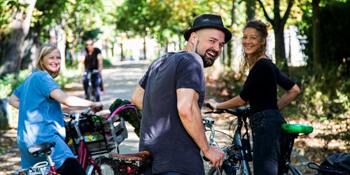 Drei Menschen stehen mit Fahrrädern auf einem sonnigen, von Bäumen gesäumten Weg und blicken lächelnd zurück in die Kamera. Im Hintergrund fährt eine weitere Person Fahrrad.