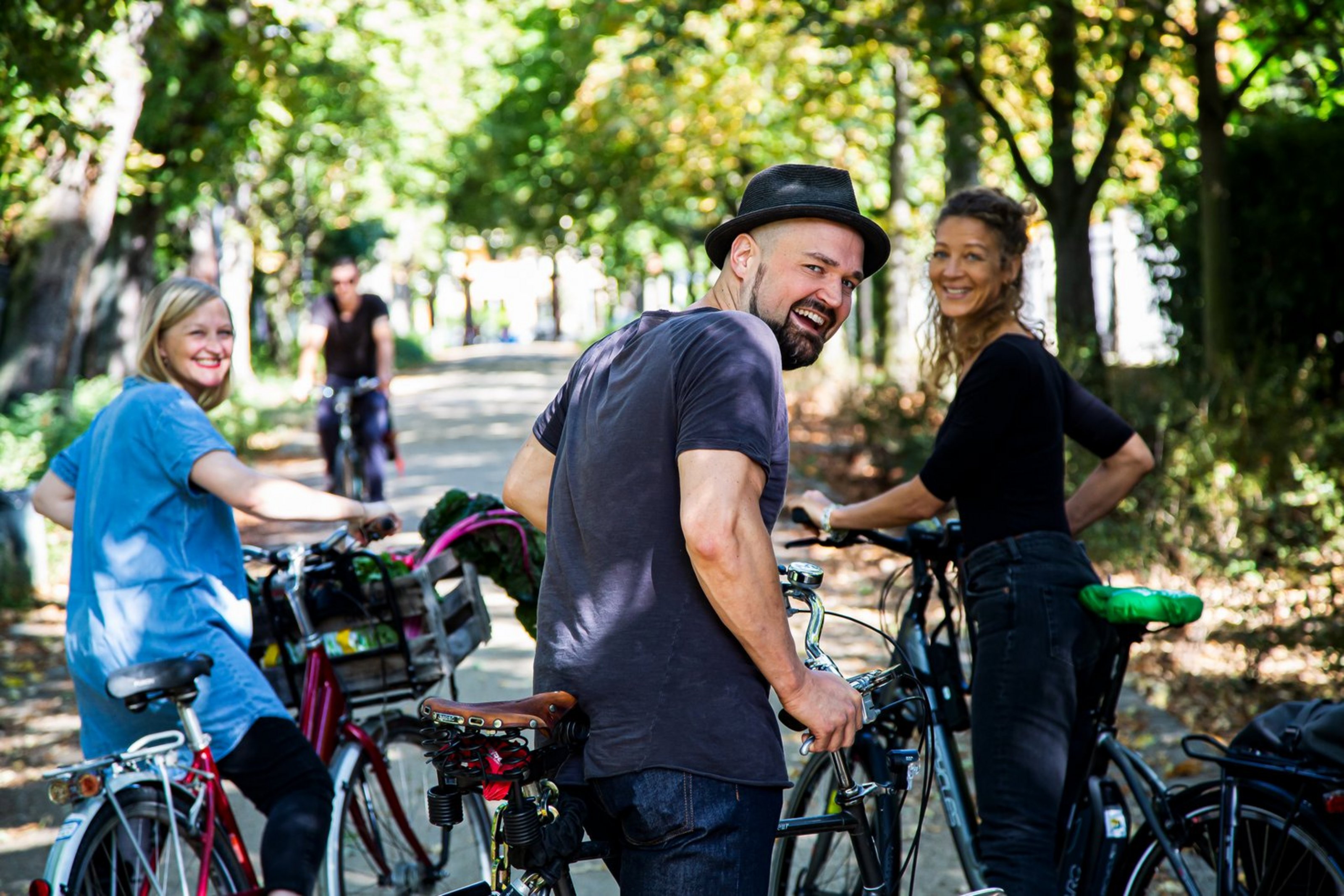 Drei Menschen stehen mit Fahrrädern auf einem sonnigen, von Bäumen gesäumten Weg und blicken lächelnd zurück in die Kamera. Im Hintergrund fährt eine weitere Person Fahrrad.
