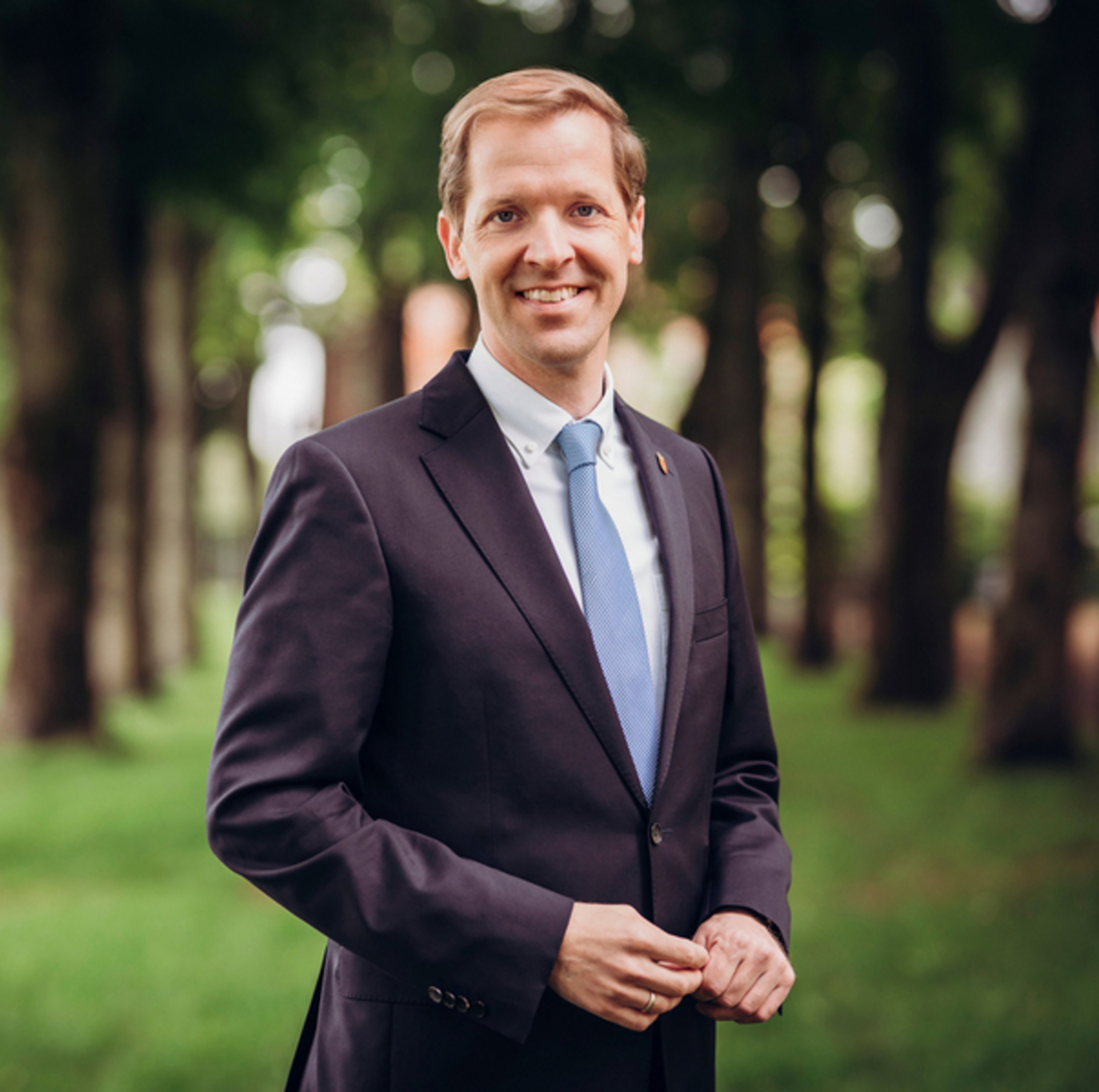 Foto: Landrat Dr. Christian Schulze Pellengahr informiert am 18. Juni über seine Aufgaben als Landrat und die Arbeit der Kreisverwaltung (Foto: Anja Tiwisina) Ein Mann steht im Freien in einem Park mit Bäumen im Hintergrund. Er trägt einen schwarzen Anzug mit einem hellblauen Hemd und einer hellblauen Krawatte. Der Mann lächelt freundlich in die Kamera und hält die Hände vor dem Körper. Die Umgebung ist grün und lichtdurchflutet.