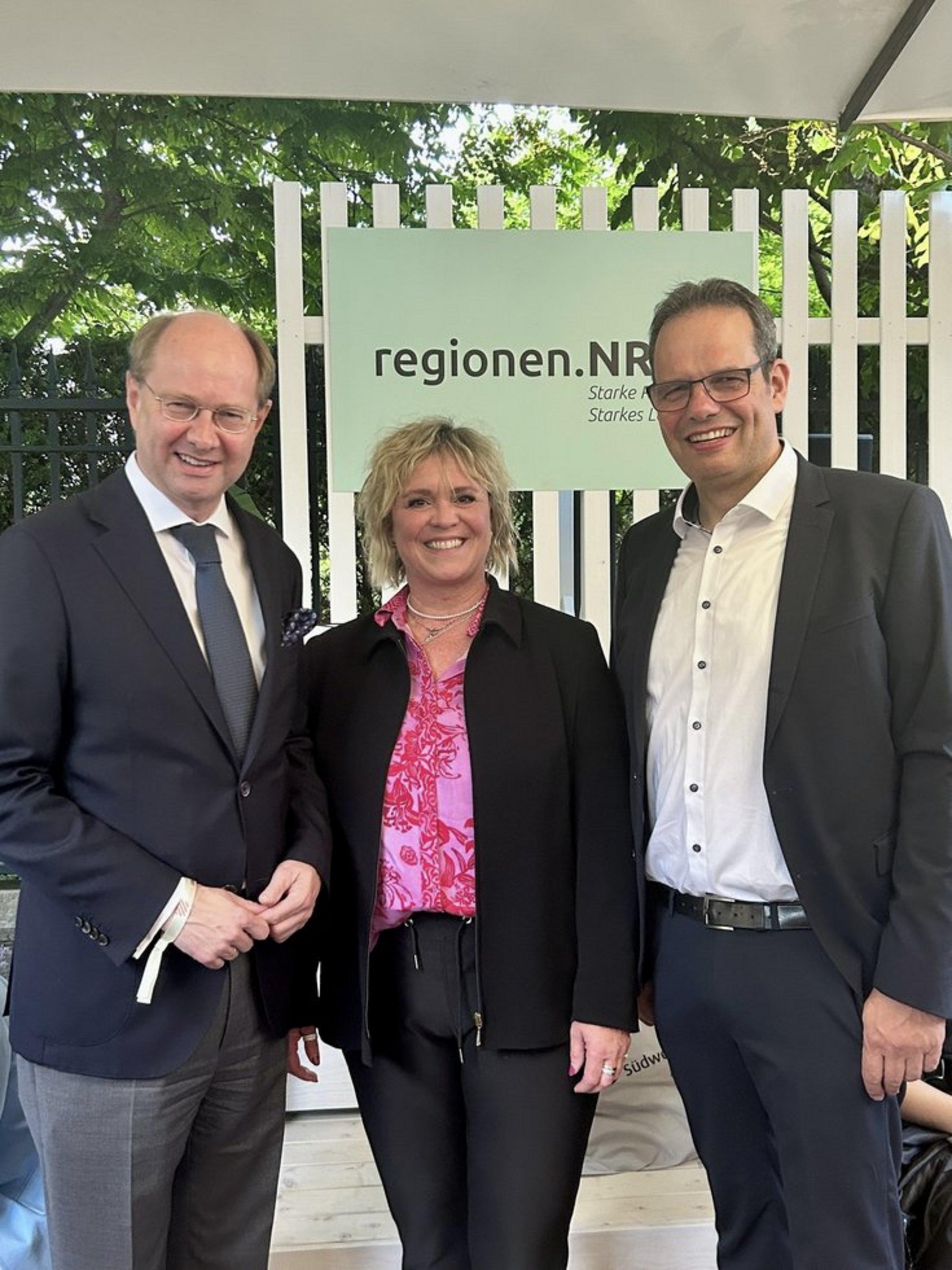 Drei Personen stehen lächelnd vor einem Schild mit der Aufschrift „regionen.NRW“.