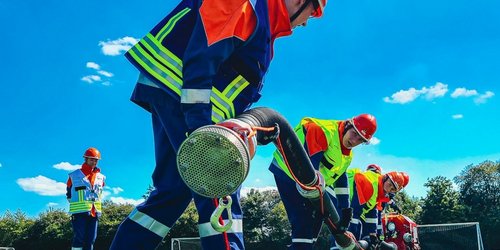 Jugendliche in Feuerwehrmontur halten einen langen Schlauch und befinden sich auf einem Fußballplatz.