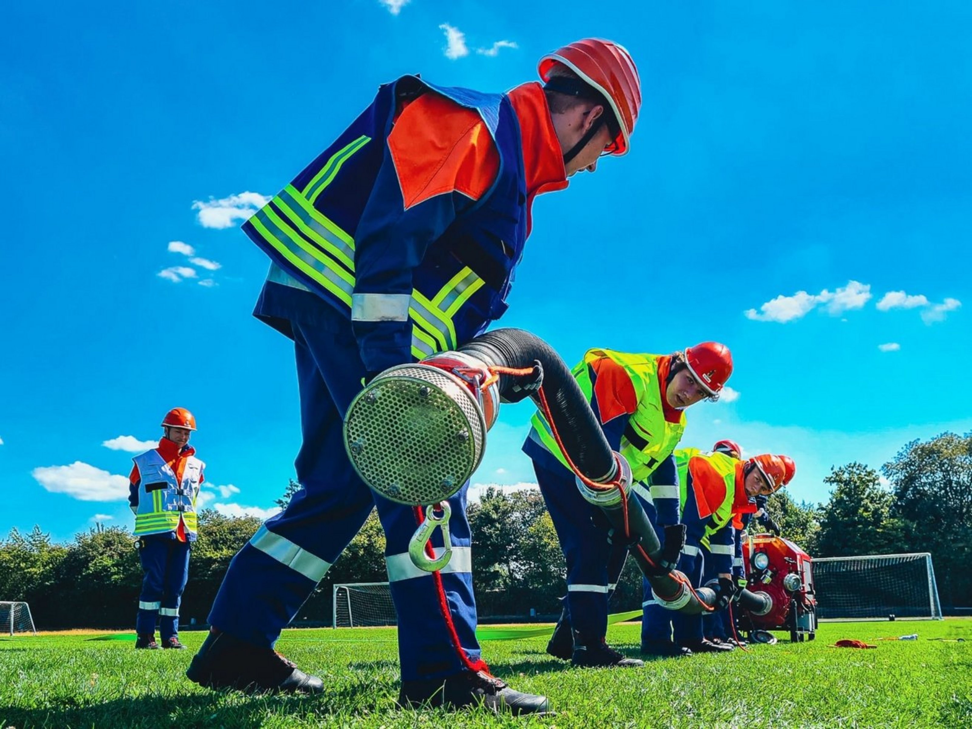 Jugendliche in Feuerwehrmontur halten einen langen Schlauch und befinden sich auf einem Fußballplatz.