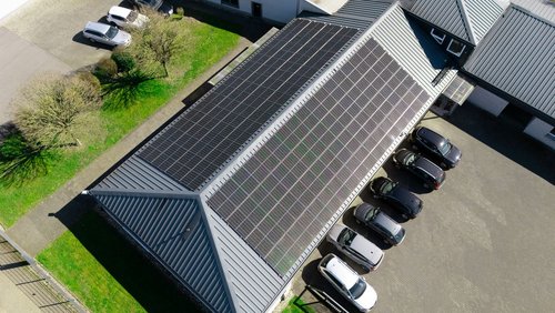 Von oben ist ein Dach mit Solaranlage zu sehen. Vor dem Haus parken mehrere Autos.