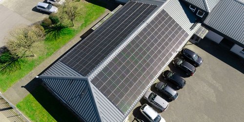 Von oben ist ein Dach mit Solaranlage zu sehen. Vor dem Haus parken mehrere Autos.