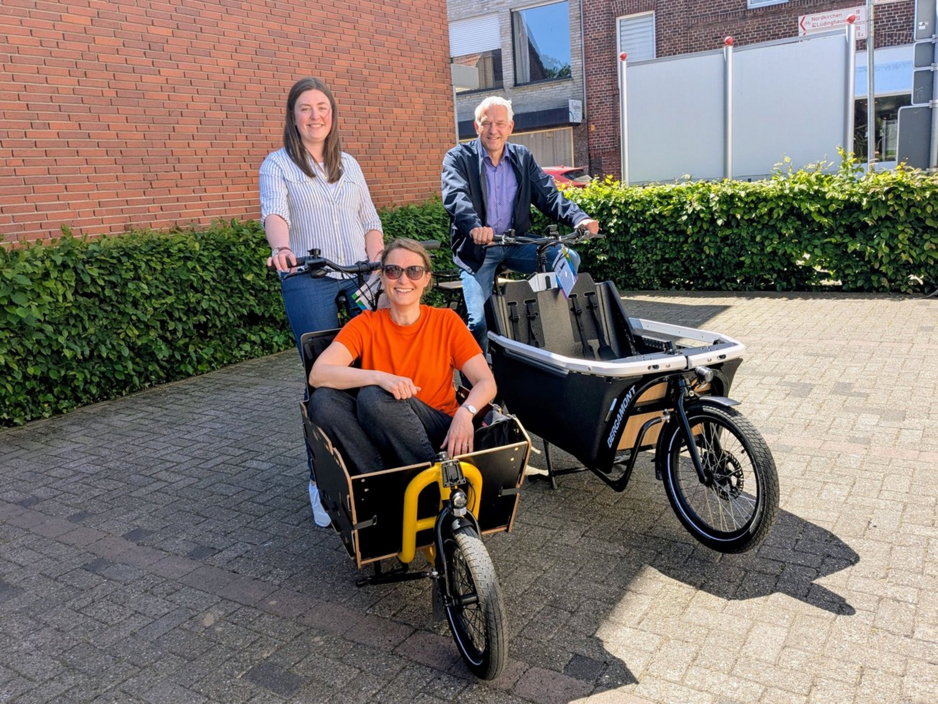 Eine Frau und ein Mann sitzen auf jeweils einem Lastenrad, die auf einem Parkplatz stehen. Auf der Ladefläche eines Lastenrades sitzt eine weitere Frau.
