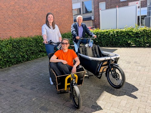 Eine Frau und ein Mann sitzen auf jeweils einem Lastenrad, die auf einem Parkplatz stehen. Auf der Ladefläche eines Lastenrades sitzt eine weitere Frau.