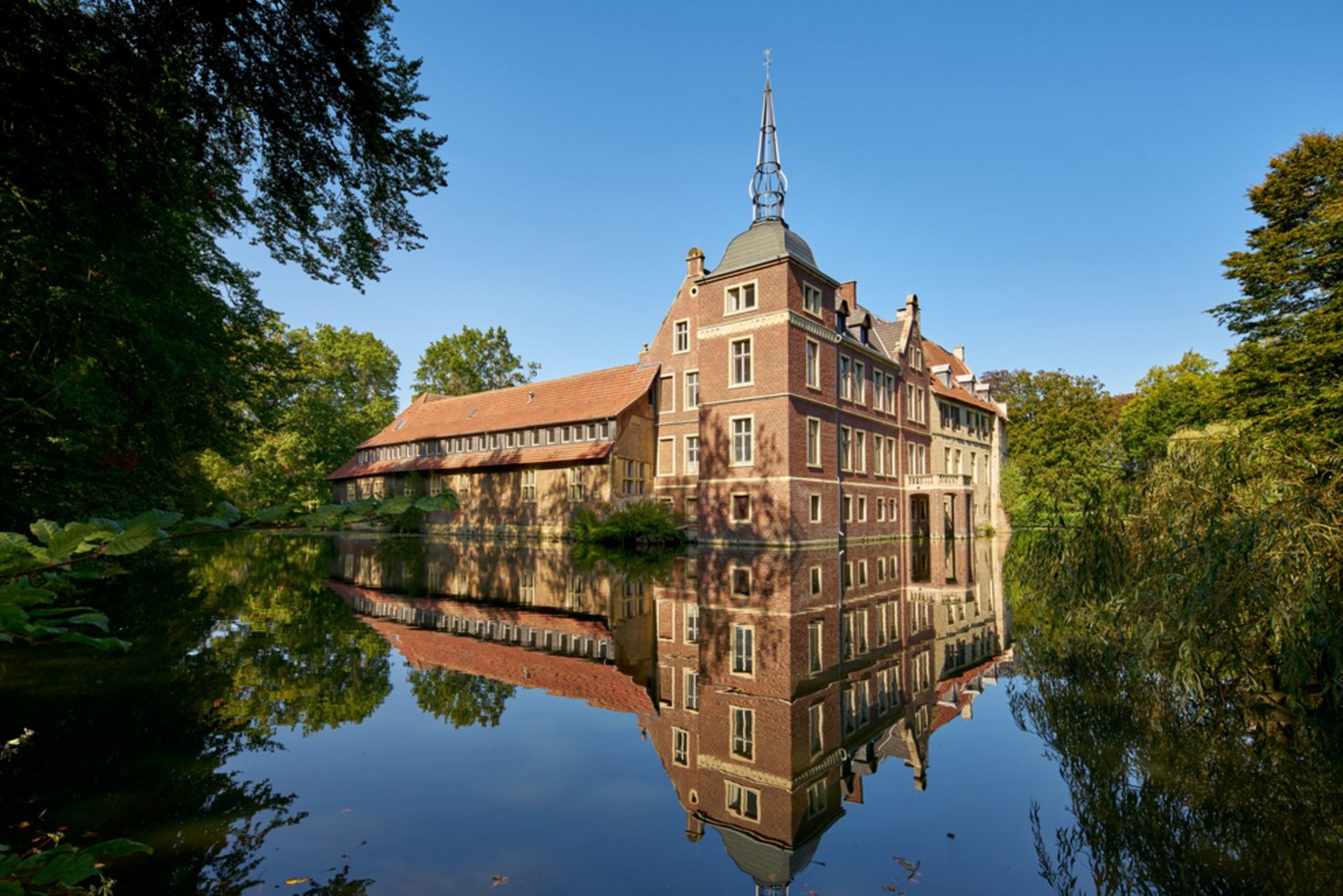 Ein Wasserschloss aus rotem Backstein ist umgeben von einem Wassergraben und Bäumen. Im Wasser spiegelt sich das Bild der Burg.