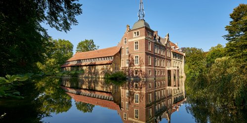 Ein Wasserschloss aus rotem Backstein ist umgeben von einem Wassergraben und Bäumen. Im Wasser spiegelt sich das Bild der Burg.