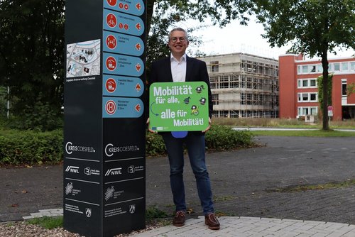 Ein Mann hält ein Plakat zur Europäischen Mobilitätswoche in Händen und steht neben einem Wegweiser einer Mobilstation.