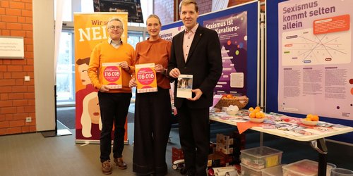 Drei Personen stehen vor Informationsständen zum Thema Gewalt- und Sexismusprävention. Von links nach rechts halten Kreisdirektor Dr. Linus Tepe und die stellvertretende Gleichstellungsbeauftragte Klaudia Müller Plakate mit der Telefonnummer des Hilfetelefons „Gewalt gegen Frauen“; rechts daneben hält Landrat Dr. Christian Schulze Pellengahr eine Broschüre.