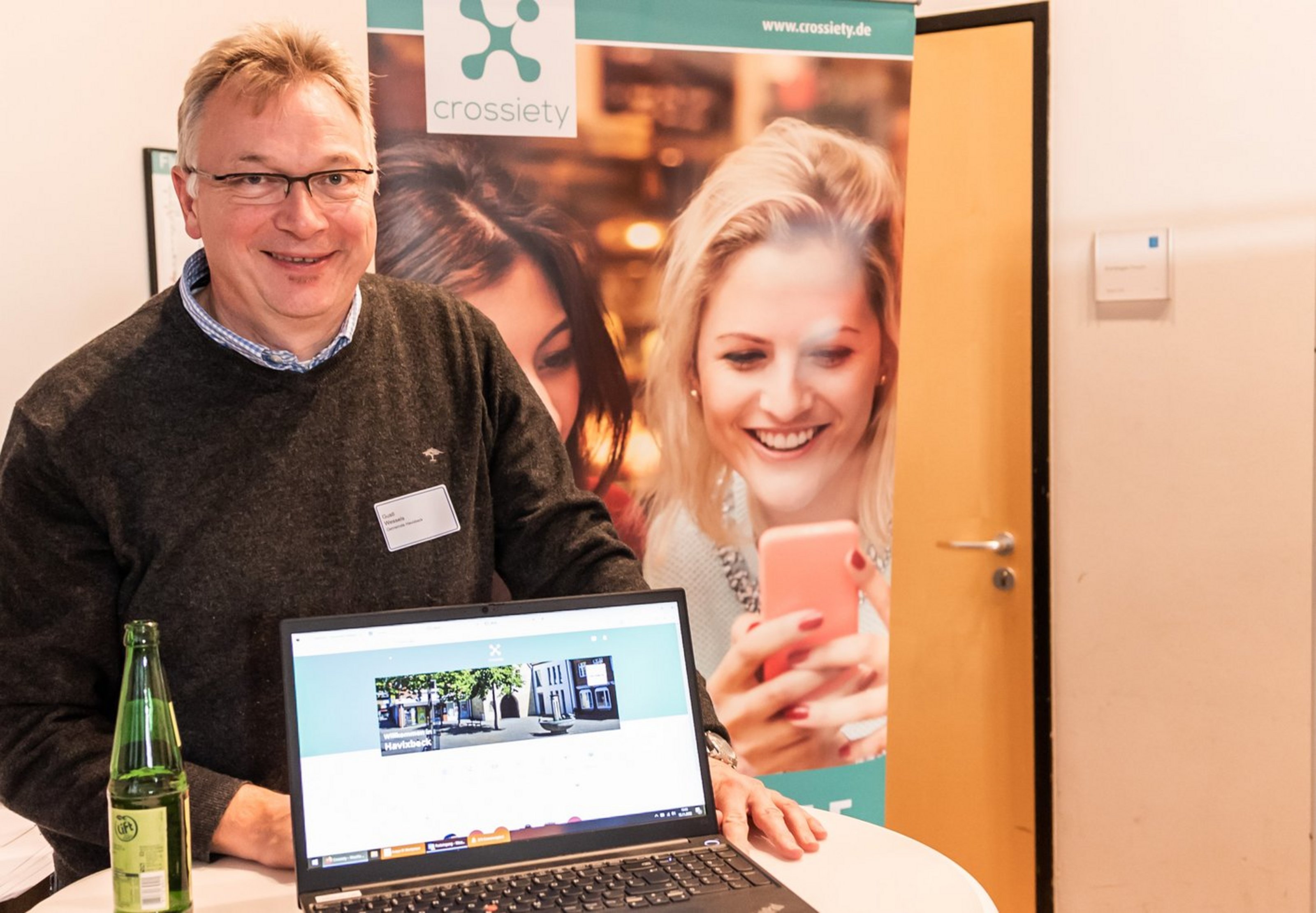 Zwei lächelnde Personen hinter einem Laptop mit geöffneter Webseite, auf einem Tisch steht eine grüne Flasche