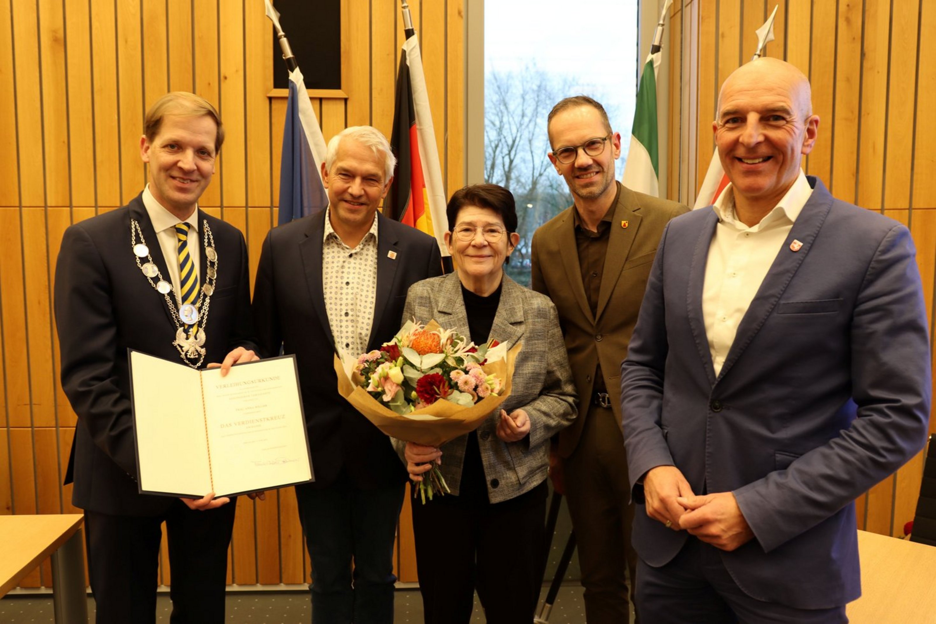 Fünf Personen stehen nebeneinander. Die Frau in der Mitte hält einen Blumenstrauß in der Hand. Der Mann links hält eine Urkunde in die Kamera.