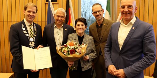 Fünf Personen stehen nebeneinander. Die Frau in der Mitte hält einen Blumenstrauß in der Hand. Der Mann links hält eine Urkunde in die Kamera.