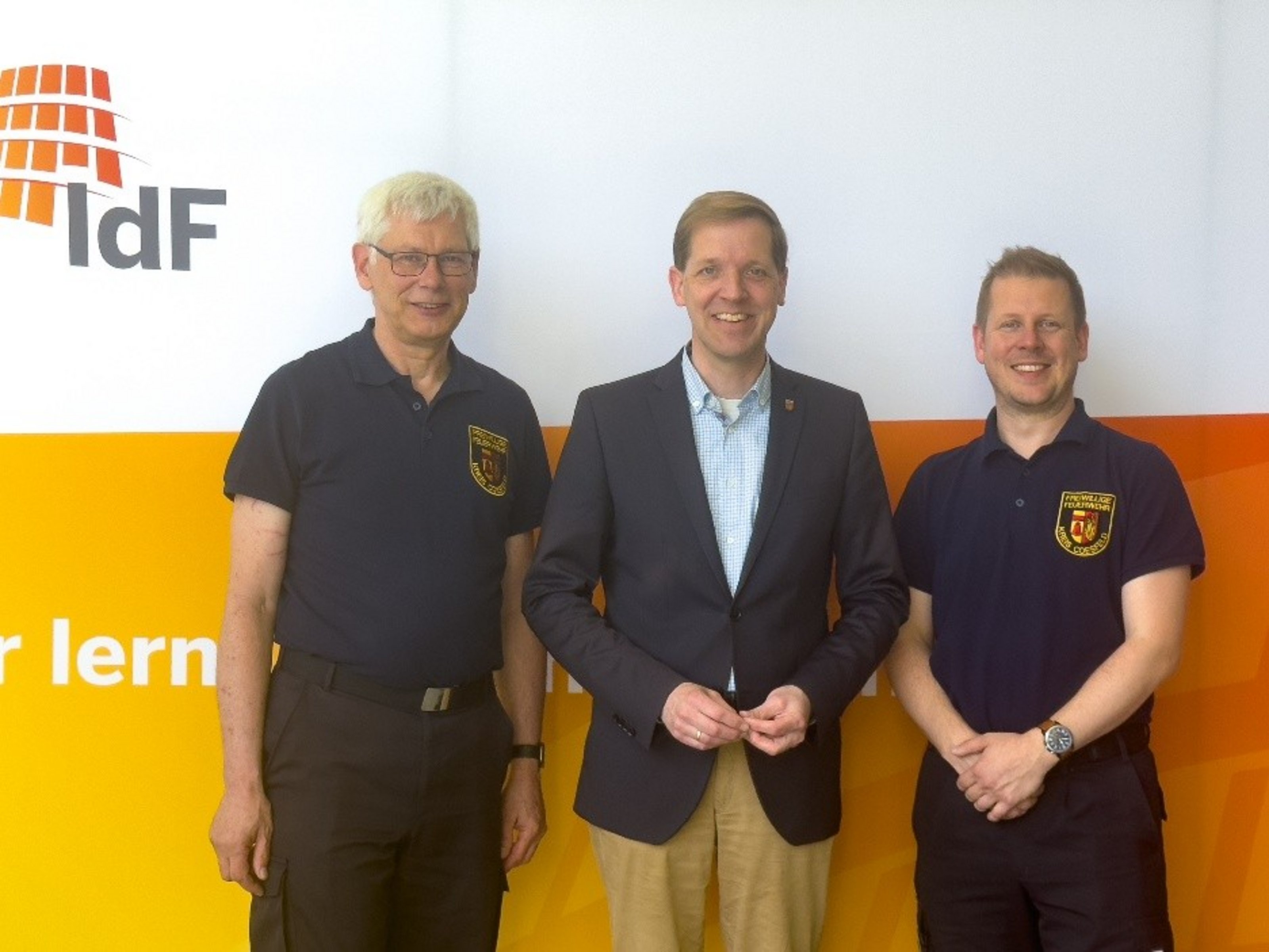 Landrat Dr. Christian Schulze Pellengahr (Mitte) mit Kreisbrandmeister Benjamin Küfer (rechts) und dem stellvertretenden Kreisbrandmeister Günter Reuver im Institut der Feuerwehr in Münster (Foto: Kreis Coesfeld). Drei Männer stehen nebeneinander und lächeln in die Kamera. Zwei von ihnen tragen dunkelblaue Polo-Shirts mit einem Feuerwehr-Emblem auf der Brust, der mittlere Mann trägt ein hellblaues Hemd und ein dunkelblaues Sakko. Im Hintergrund ist ein weiß-orangefarbener Hintergrund mit dem Logo „IdF“ zu sehen, das für das Institut der Feuerwehr steht.