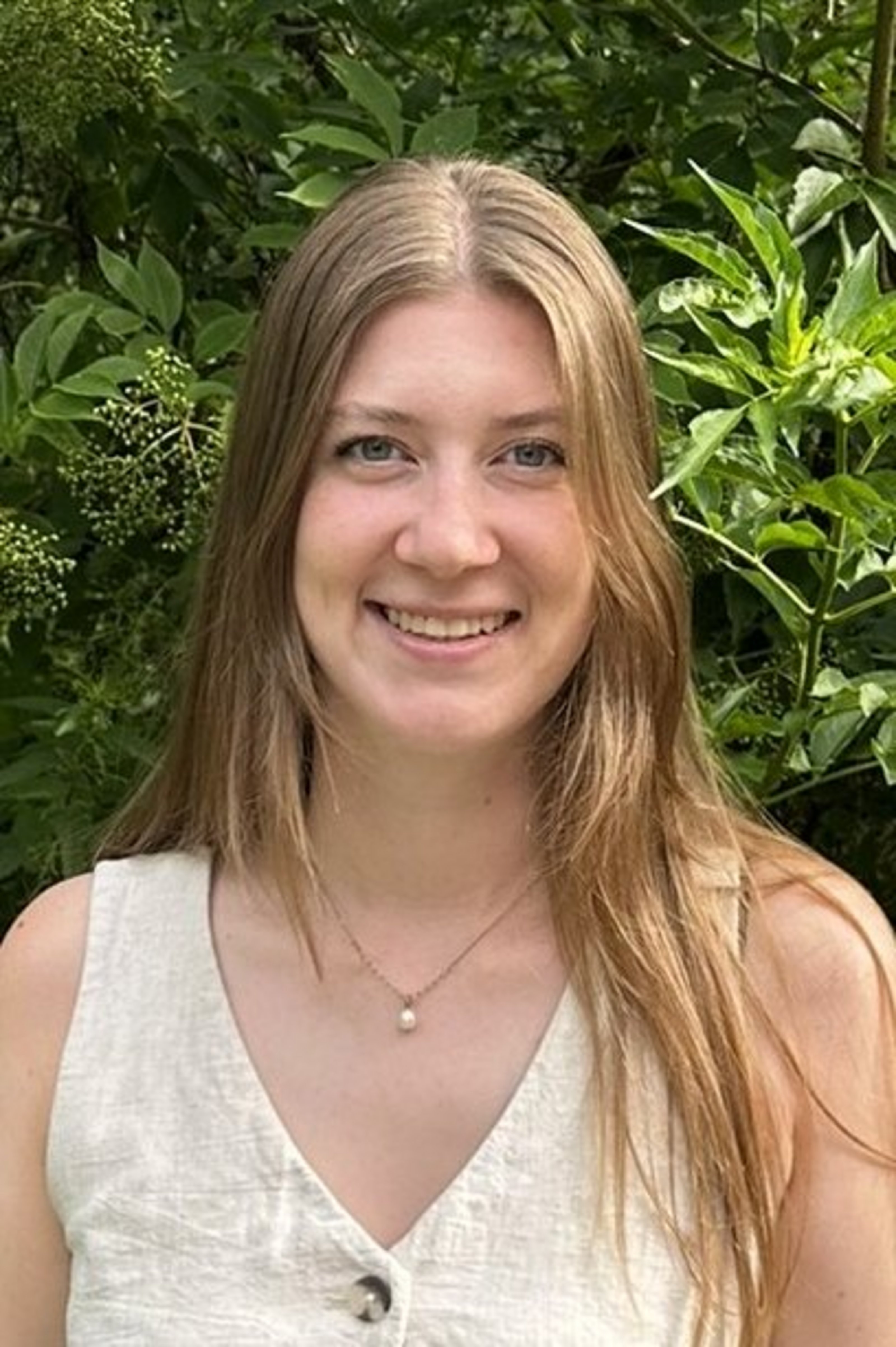 Ein Porträtbild mit der Ansprechpartnerin Nina Hünteler: Sie trägt eine blaue geblümte Bluse und steht vor einer Hecke.